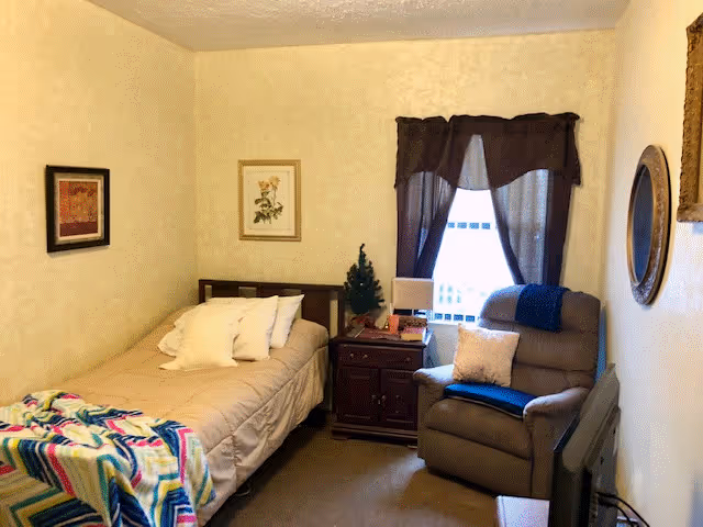 Small furnished bedroom with a single bed, nightstand, recliner chair, window with curtains, and wall art.