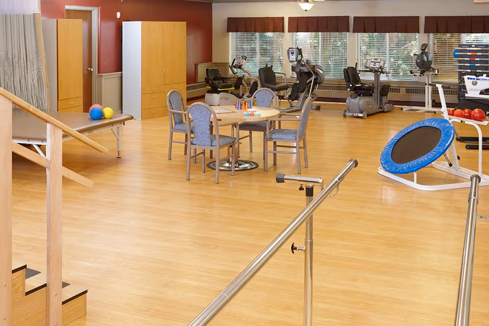 A spacious physical therapy or rehabilitation room with wooden flooring, exercise equipment including stationary bikes, a small trampoline, parallel bars, and a table with four chairs in the center. There are cabinets and a therapy table with colorful balls on it, and large windows with brown valances letting in natural light.