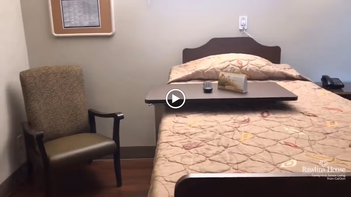 A senior living facility bedroom with a single bed covered by a patterned quilt. There is a wooden overbed table on the bed holding a remote control and a small card. To the left of the bed is a cushioned armchair with wooden armrests. The walls are plain with a framed item hanging above the chair. The floor is wooden.