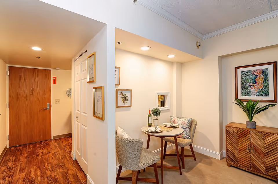Small dining area with a round table set for two, featuring a bottle of wine, two plates, and floral cushions on two chairs. The area is well-lit with recessed ceiling lights and decorated with framed botanical prints on the wall. Adjacent to the dining area is a wooden cabinet with a potted plant and a framed artwork above it. The entrance door and a closet with white double doors are visible in the background, along with wood flooring near the entrance and carpet in the dining area.