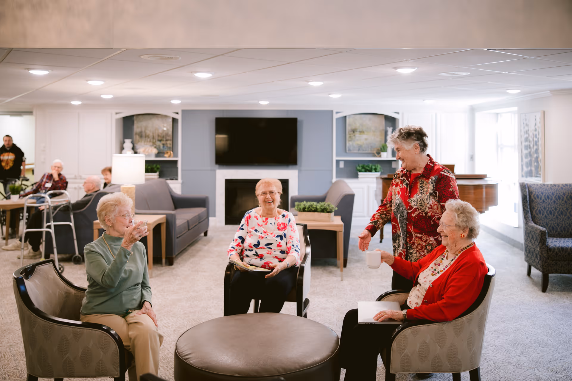 A group of elderly women sitting and standing in a bright, comfortable common area with chairs, a round ottoman, and a fireplace with a TV above it. They are smiling and engaging in conversation, with other seniors and a walker visible in the background.