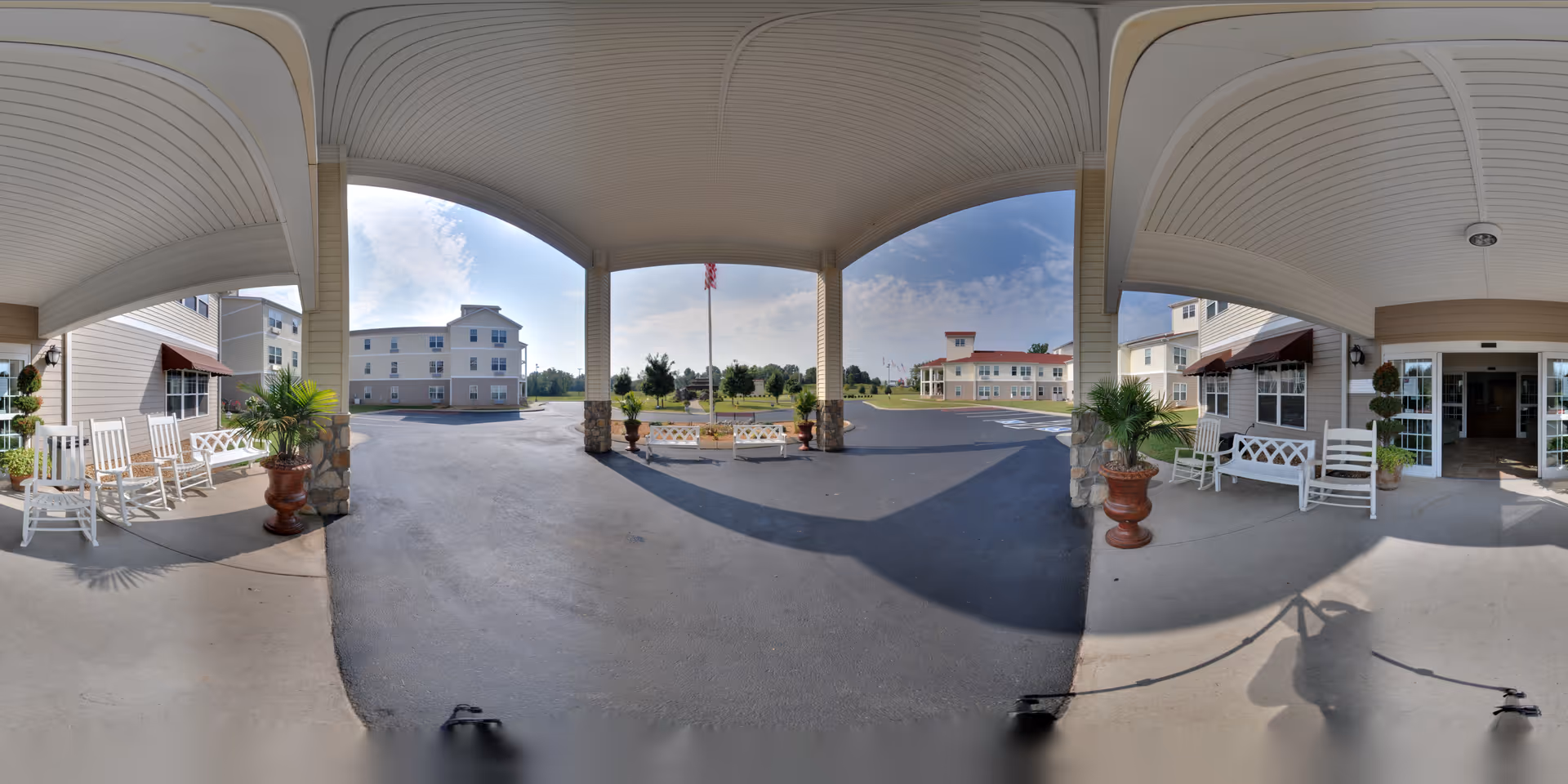 Covered entrance area of a senior living facility with white rocking chairs and benches on either side, large potted plants, and a view of the driveway and buildings in the background under a partly cloudy sky.