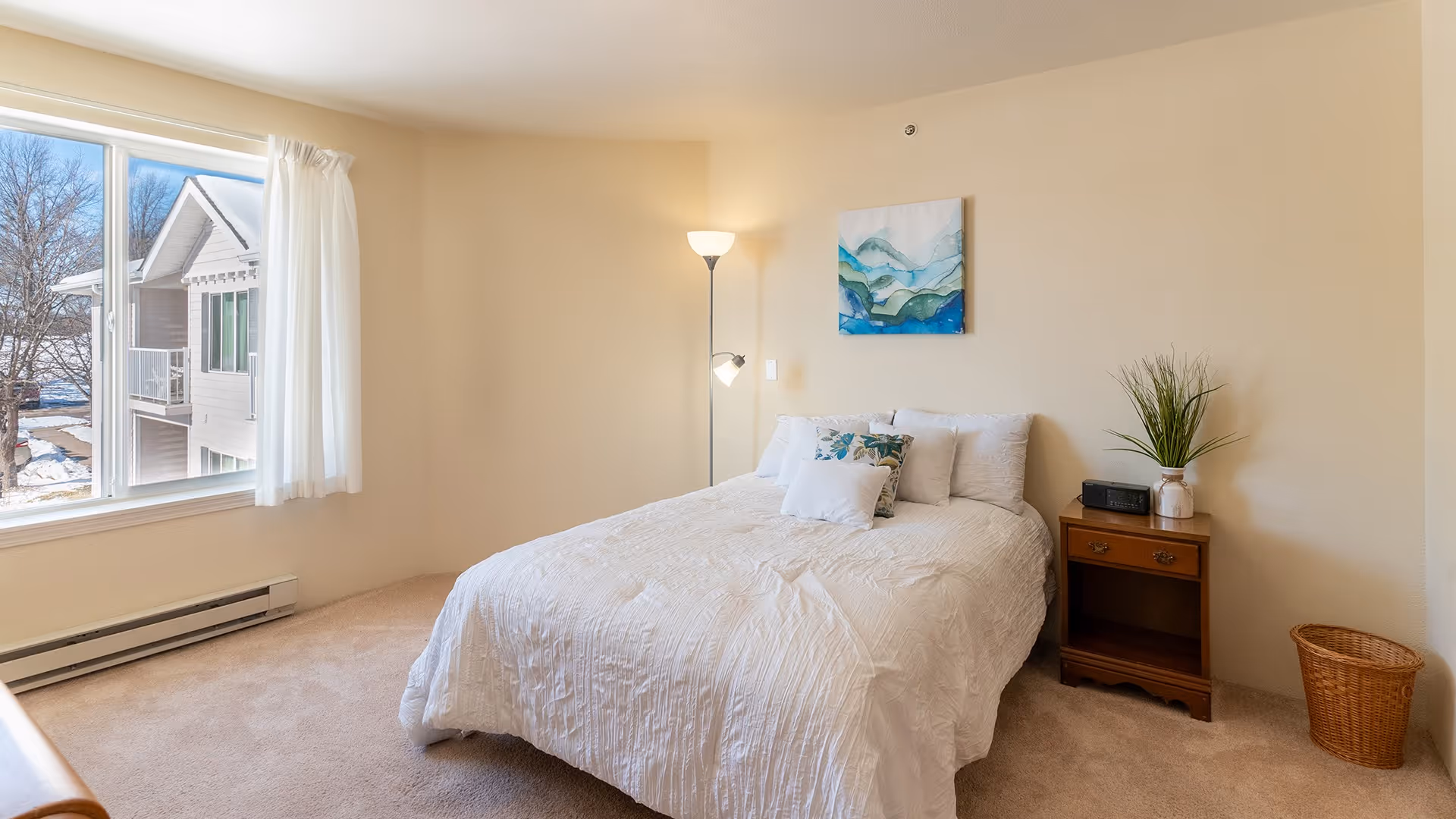A bright bedroom with a large window showing an outdoor view of a neighboring building and trees. The room features a bed with white bedding and decorative pillows, a wooden nightstand with a plant and a clock radio, a floor lamp, and a wicker wastebasket. A piece of abstract art hangs on the wall above the bed.