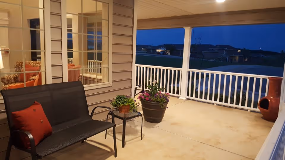 Covered outdoor patio area at dusk with a black bench featuring a red cushion, a small glass-top side table with a potted plant, a large flower pot with blooming flowers, and a clay chiminea. The patio has white railing and overlooks a grassy area with houses in the distance. Through the window, an indoor seating area with patterned armchairs and an orange cushion is visible.