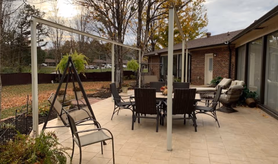 Outdoor patio area with a tiled floor featuring a round dining table surrounded by six chairs, two cushioned lounge chairs, and a swing chair. The patio is adjacent to a brick building with large windows and a door. Trees with autumn leaves and a grassy yard are visible in the background.