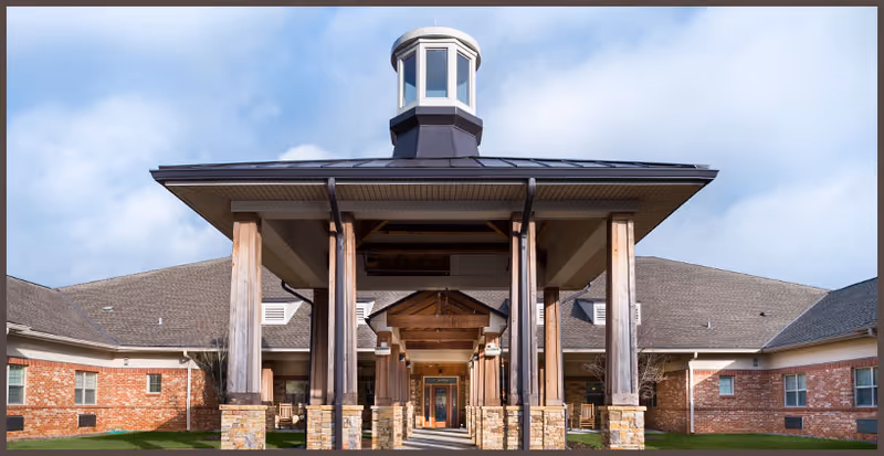 Front exterior view of a single-story brick building with a covered entrance supported by wooden pillars and a small cupola on the roof under a partly cloudy sky.