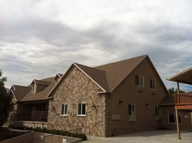 Exterior view of a senior living facility building with a stone facade on one side and beige siding on the other, multiple windows, and a sloped roof under a cloudy sky.