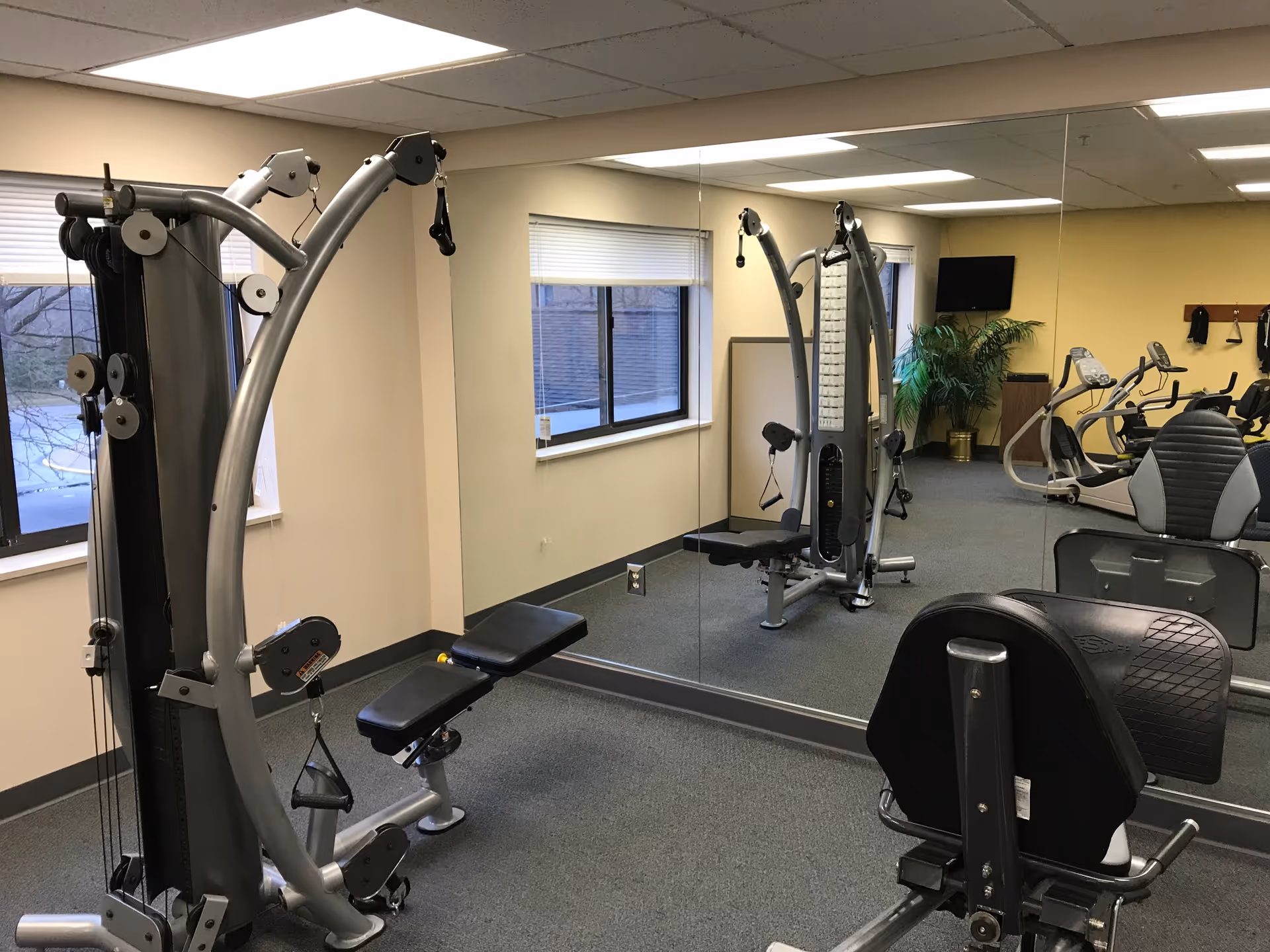 A fitness room with exercise equipment including a multi-function weight machine, stationary bikes, and a large wall mirror reflecting the room. The room has beige walls, windows with blinds, and a potted plant in the corner.