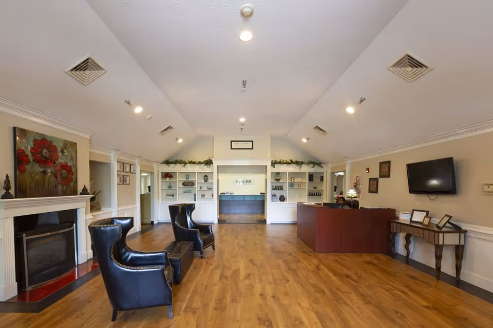 A spacious reception area of Rock Hill Post Acute Care Center featuring a wooden floor, two black leather armchairs with a small table between them, a fireplace with a floral painting above it on the left, a wooden reception desk on the right, a wall-mounted TV, and built-in white shelves with decorative items and greenery. The ceiling has recessed lighting and air vents.
