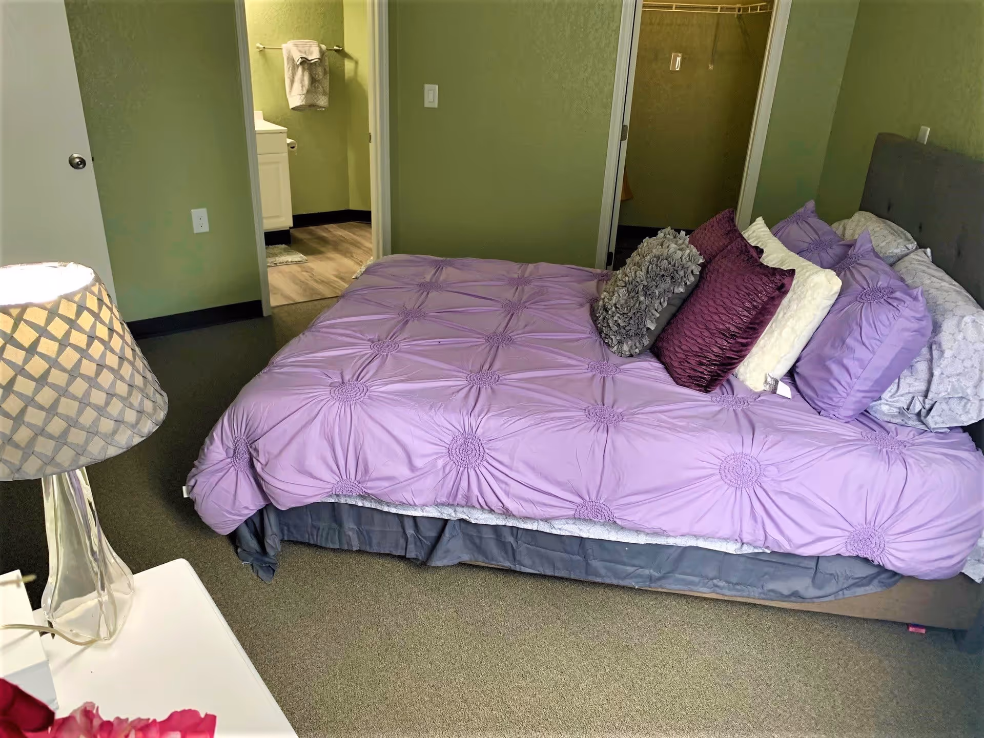 A bedroom with a bed covered in a purple comforter and multiple pillows in shades of purple, white, and gray. A bedside table with a clear lamp and a pink flower is visible in the foreground. The walls are painted green, and there is an open door leading to a bathroom with a towel hanging on the wall.