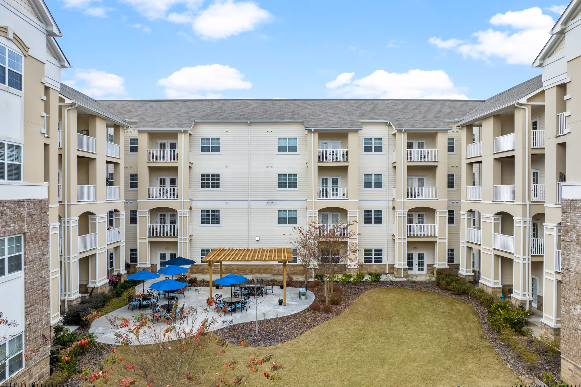 Outdoor courtyard area of a senior living facility with a pergola, several tables with blue umbrellas, surrounded by a multi-story beige building with balconies and windows under a partly cloudy sky.