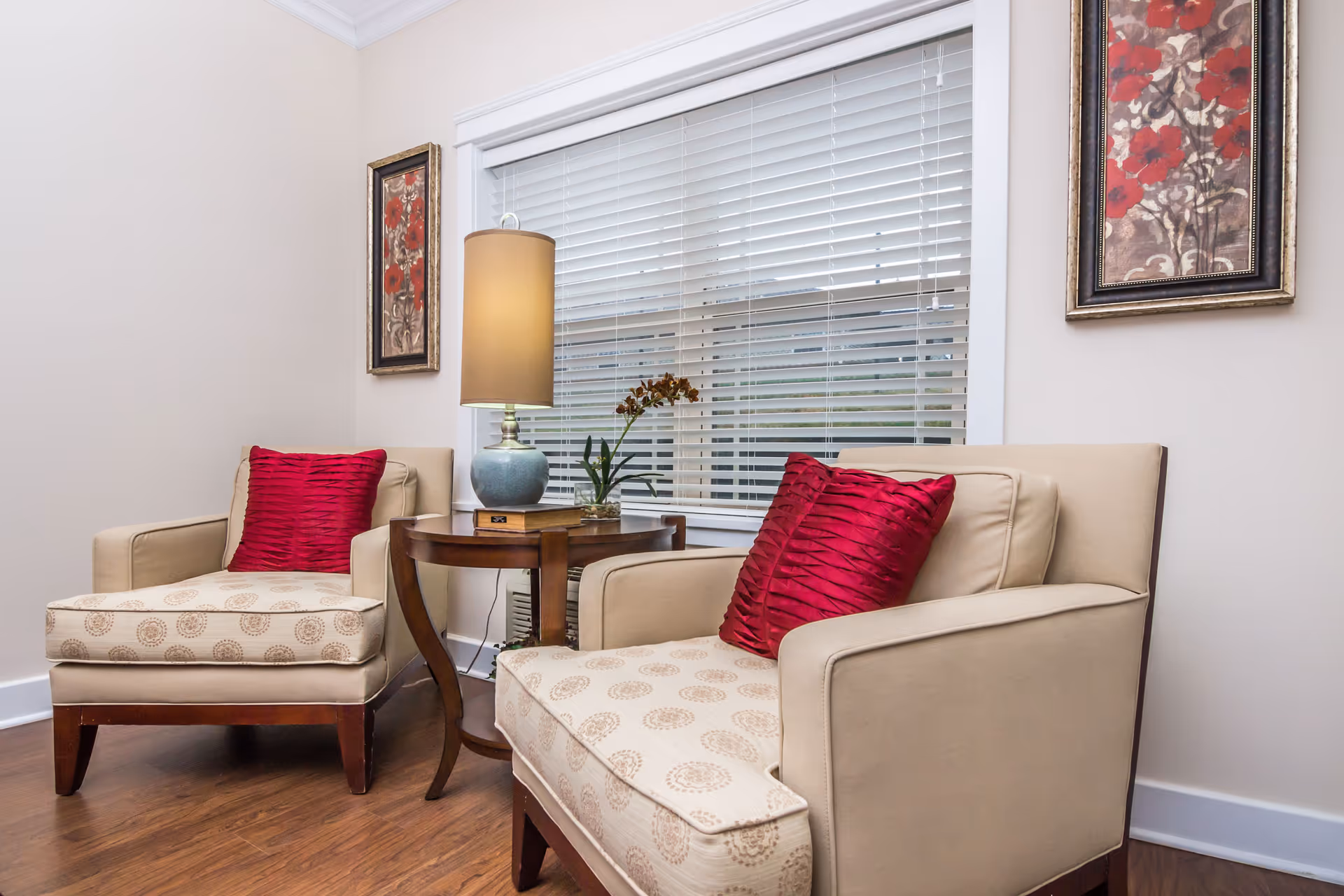 A cozy sitting area with two beige armchairs featuring patterned cushions and red throw pillows, separated by a wooden side table with a lamp and a small plant. The background includes a window with white blinds and two framed floral artworks on the wall.