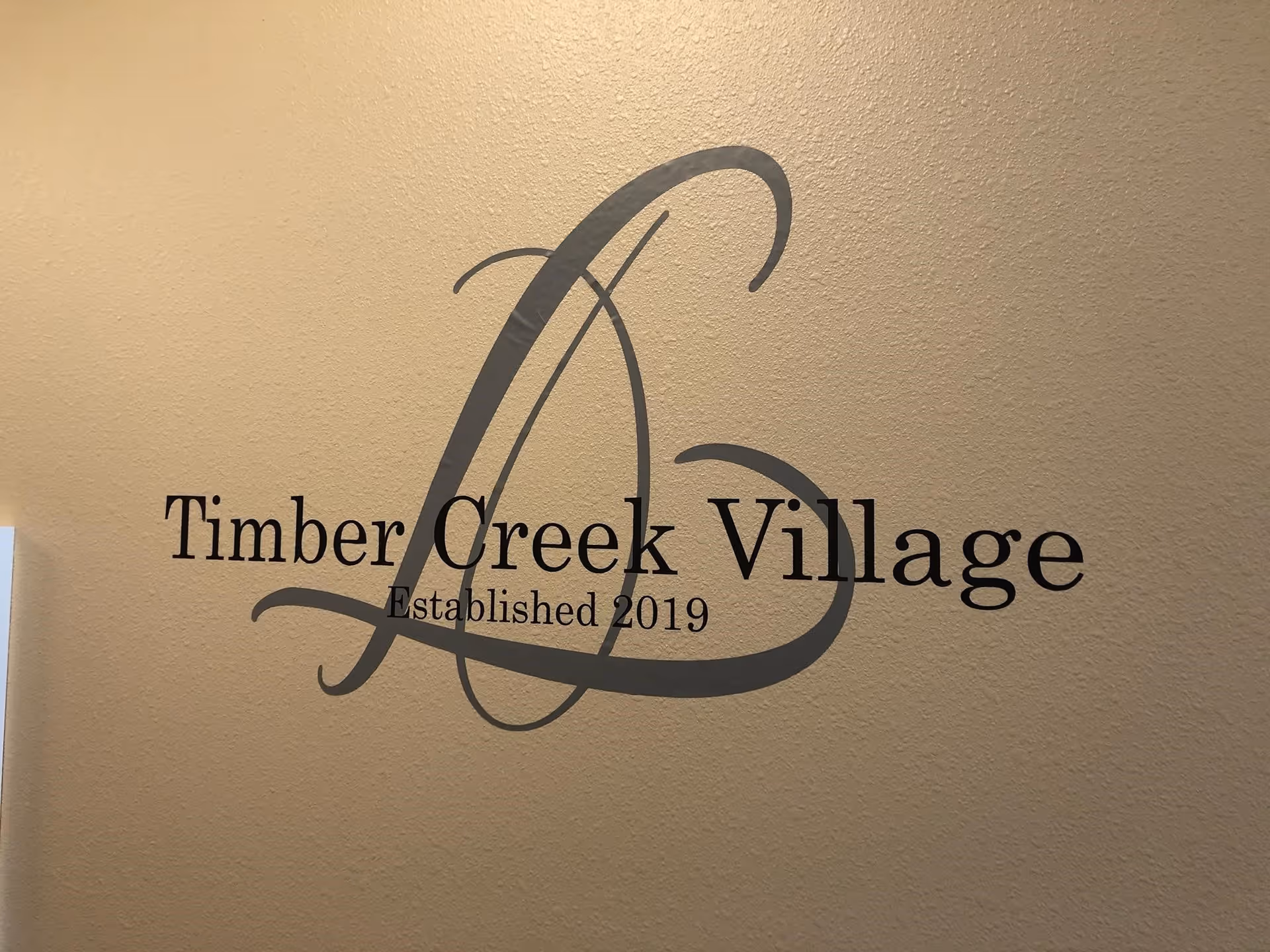 A decorative wall sign that reads "Timber Creek Village Established 2019" on a textured beige interior wall.