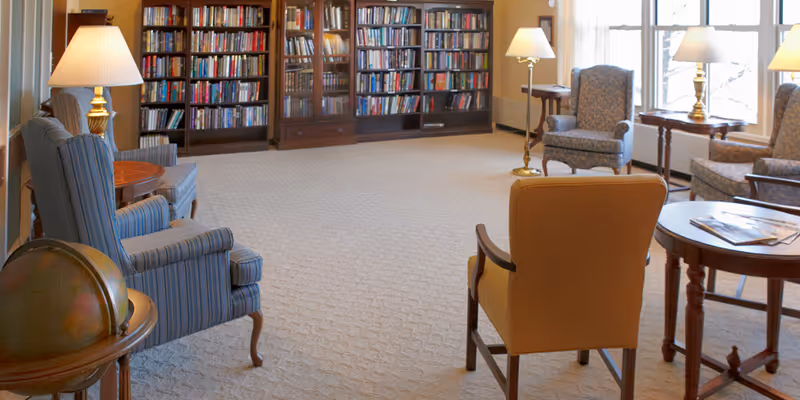 A cozy senior living common area with several upholstered armchairs arranged around small wooden tables. There are floor lamps providing warm lighting, large windows letting in natural light, and a large bookshelf filled with books along the back wall. A globe sits on a small table in the foreground.