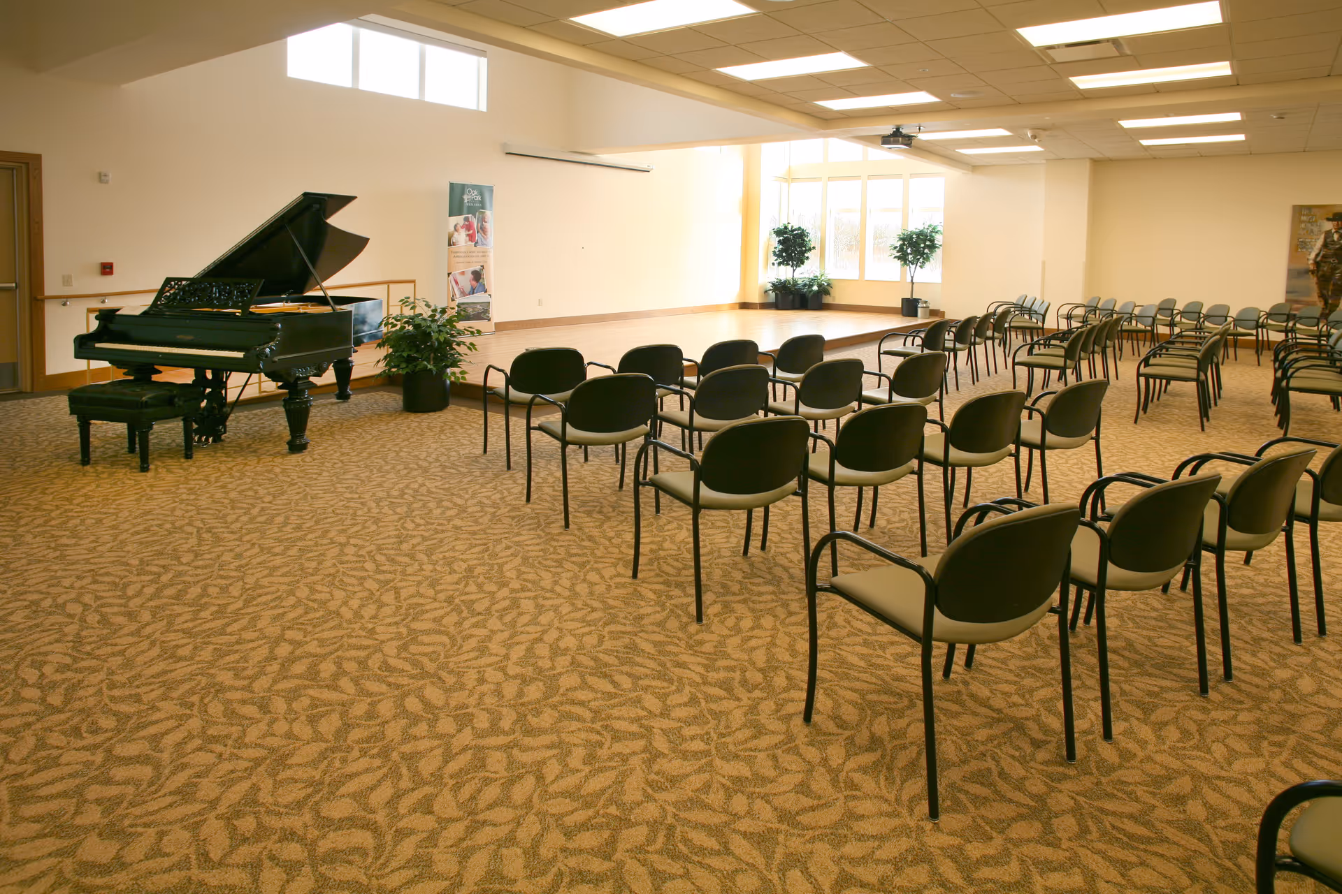 Spacious multipurpose room with rows of chairs facing a small stage and a grand piano.