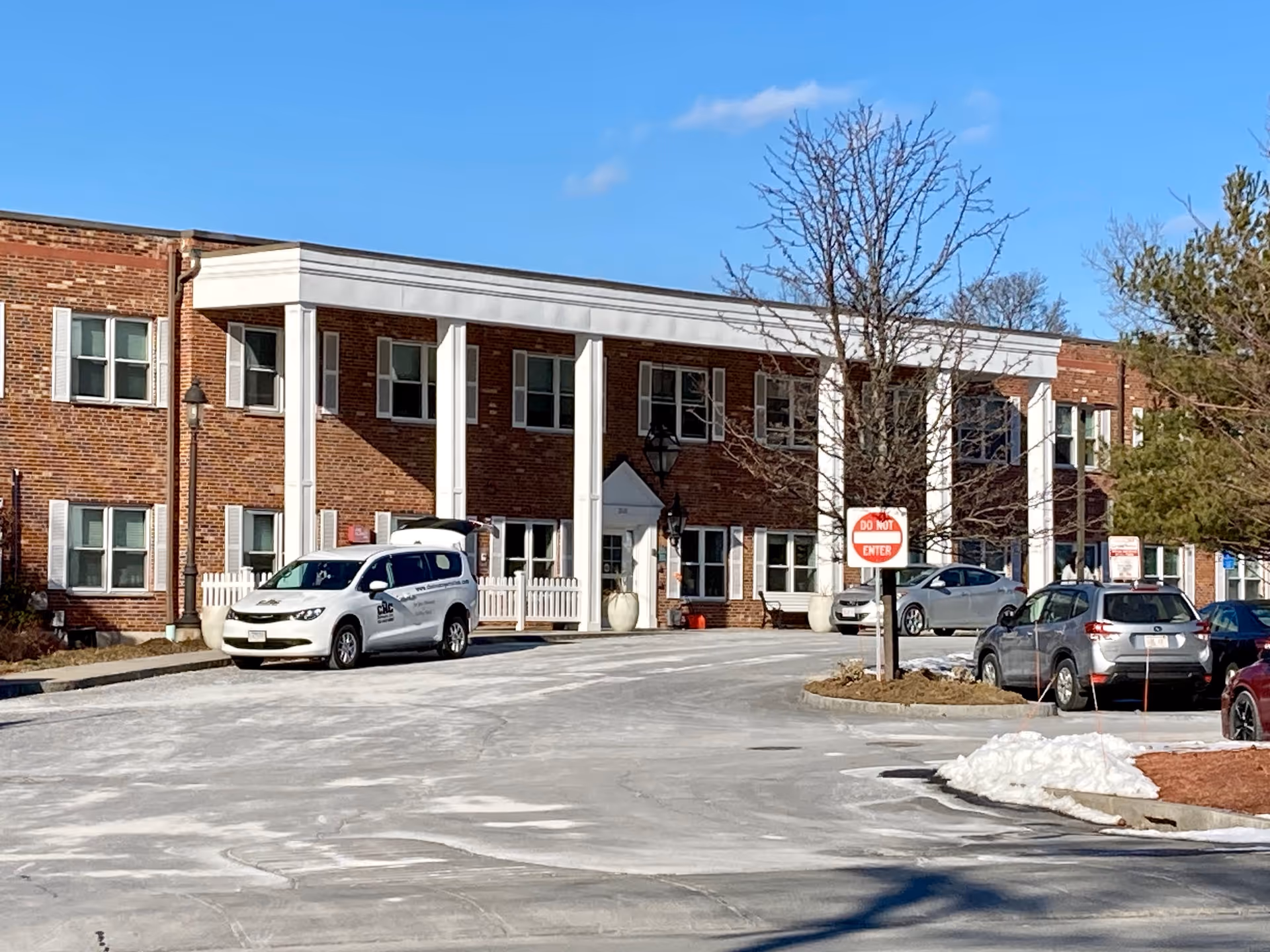 Brick two-story senior living facility front with white columns, parked cars, and a 'Do Not Enter' sign.