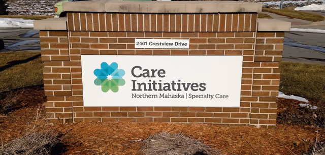 A brick signpost with a white sign displaying the logo and text 'Care Initiatives Northern Mahaska | Specialty Care' and an address plaque above it reading '2401 Crestview Drive'. The sign is outdoors with some patches of snow and grass visible around it.