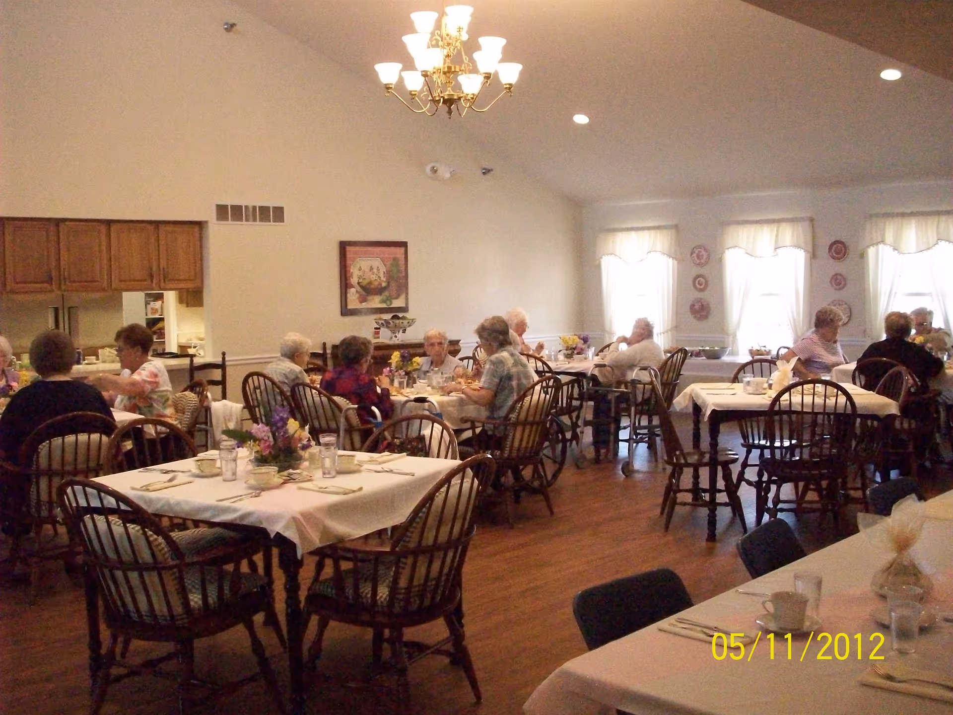 A dining room in a senior living facility with several elderly people seated at tables covered with white tablecloths. The room has wooden chairs, a chandelier hanging from the ceiling, and windows with white curtains letting in natural light. There is a kitchen pass-through window with wooden cabinets visible on the left side.