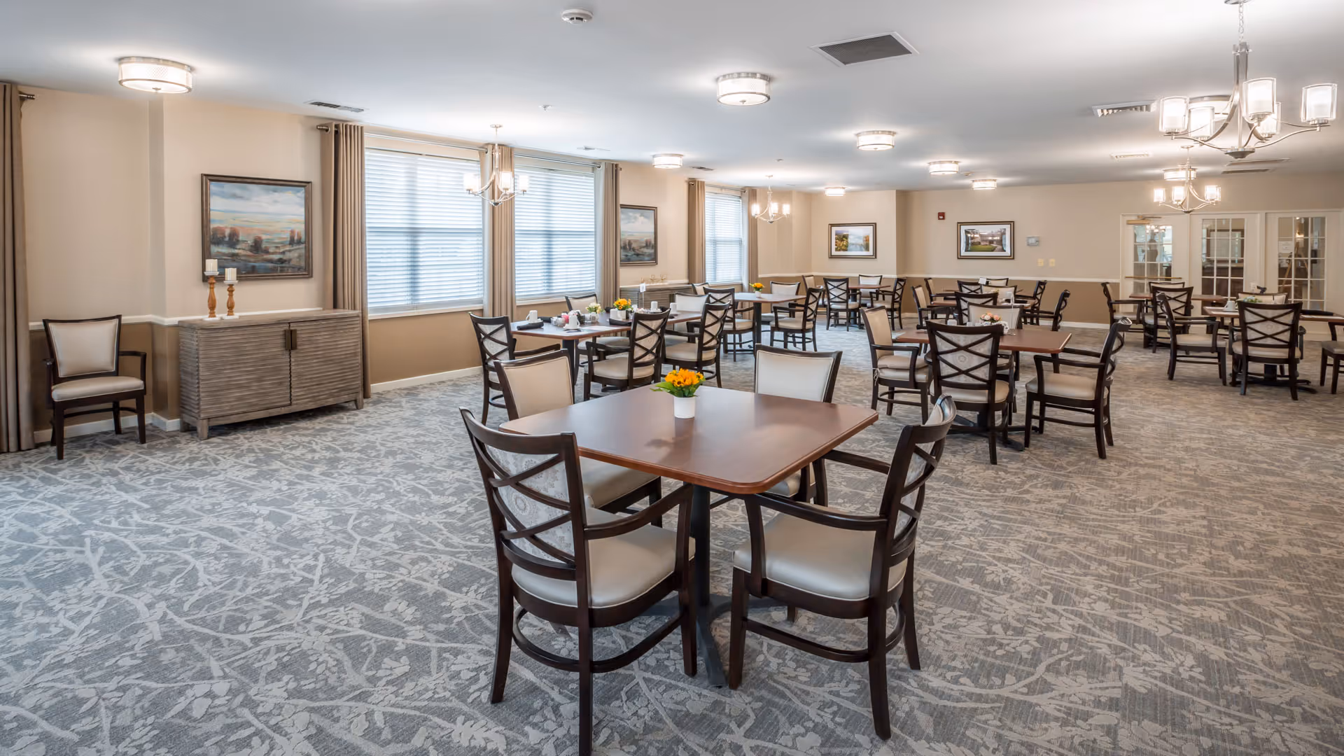A spacious dining room with multiple wooden tables and chairs arranged neatly. The room has large windows with blinds, beige walls adorned with framed paintings, and modern ceiling light fixtures. The carpet has a subtle leaf pattern, and small flower arrangements are placed on the tables.