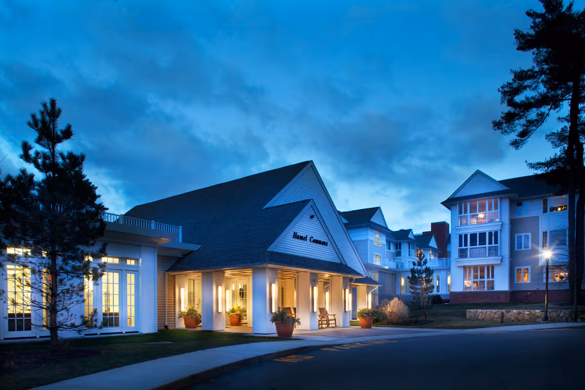 Exterior view of Fuller Village senior living facility at dusk, showing a well-lit entrance with potted plants and a driveway, surrounded by multi-story residential buildings and trees under a cloudy blue sky.