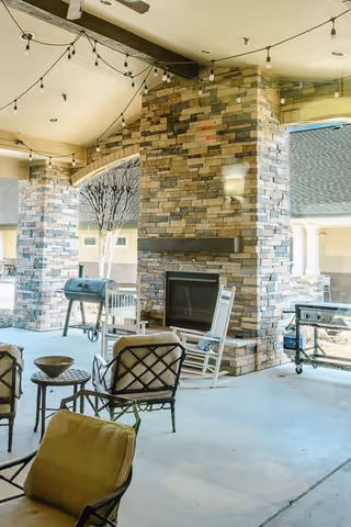 Covered outdoor seating area with cushioned chairs and a rocking chair arranged around a stone fireplace. String lights hang from the ceiling, and there is a barbecue grill and a small table nearby. The area is open to the outside with stone pillars supporting the roof.