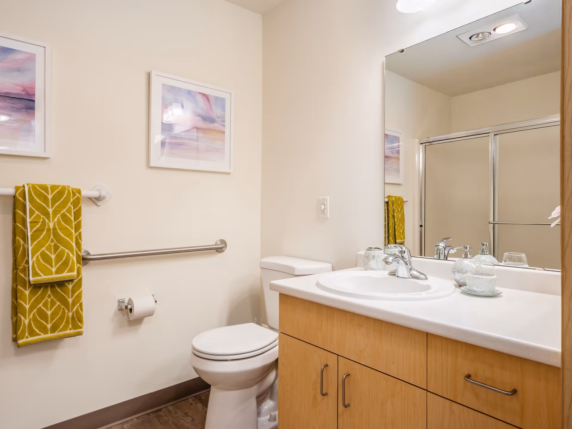 A clean bathroom with a white toilet, a wooden vanity with a white countertop and sink, a large mirror above the sink, a glass soap dispenser, and a glass container on the counter. There is a towel rack on the wall with two yellow towels featuring a leaf pattern, a toilet paper holder with a roll of toilet paper, and a framed watercolor artwork hanging above the towel rack. A shower with sliding glass doors is visible in the mirror reflection.