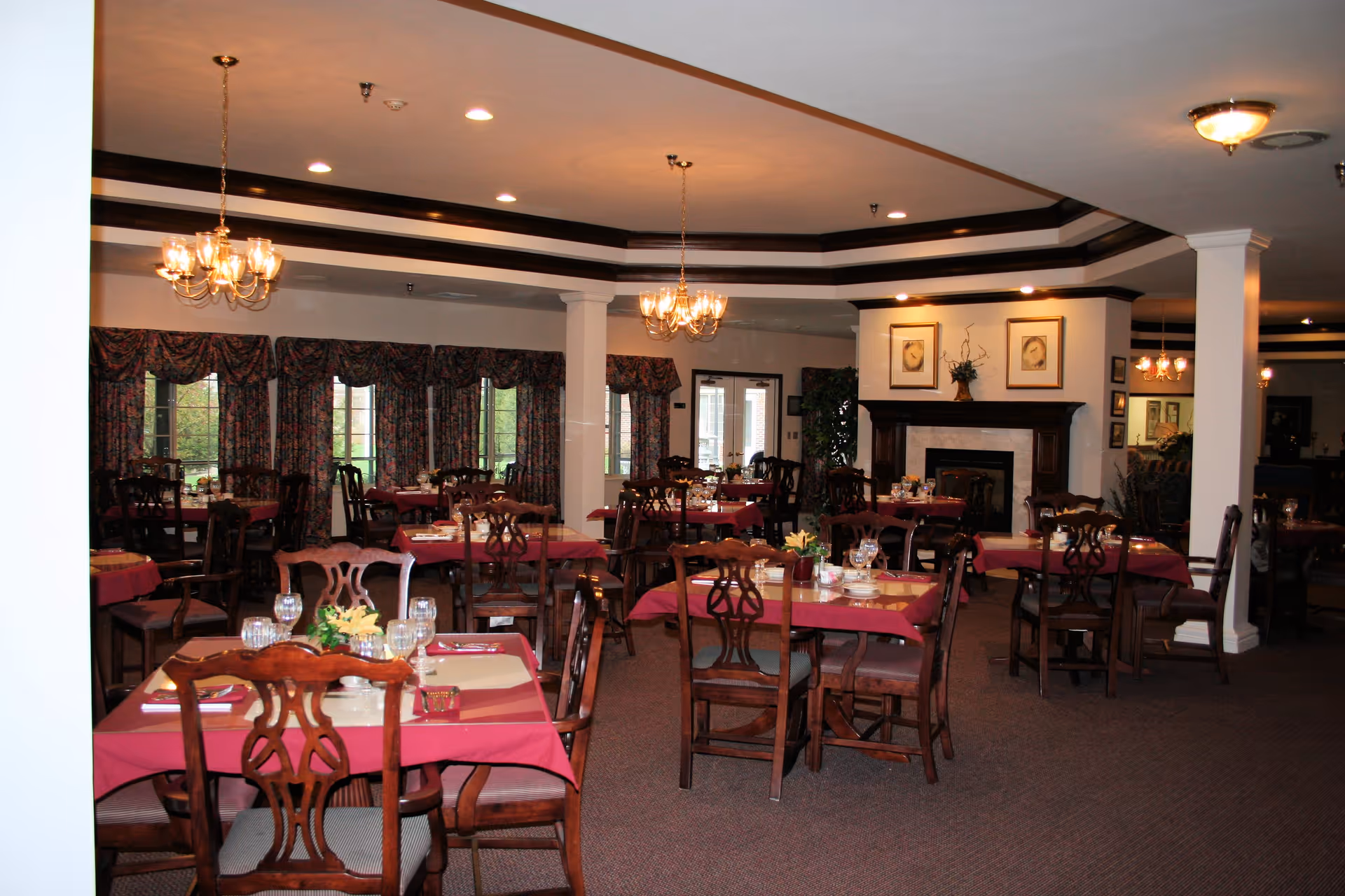 A dining room with multiple wooden tables and chairs arranged neatly. Each table is covered with a red tablecloth and set with glassware, napkins, and small floral centerpieces. The room features chandeliers hanging from the ceiling, large windows with patterned curtains, and a fireplace with framed artwork above it.