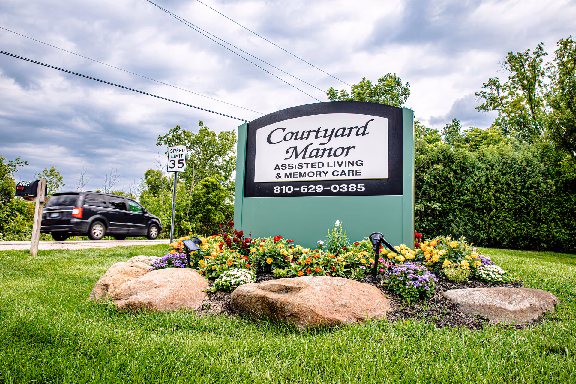 A large green and black sign for Courtyard Manor Assisted Living & Memory Care surrounded by colorful flowers and rocks, with a speed limit 35 sign and a black vehicle driving on a road in the background under a cloudy sky.