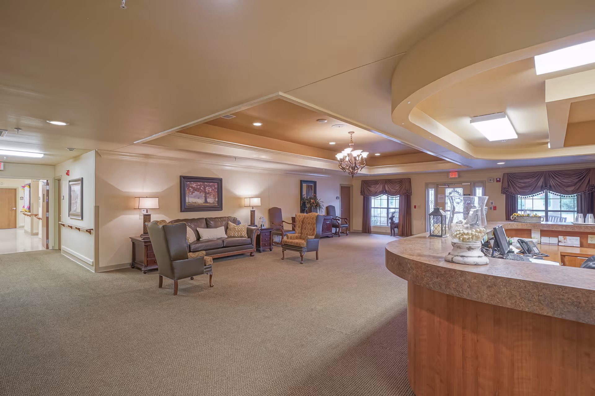 A spacious and well-lit senior living facility common area with comfortable seating including armchairs and a sofa, side tables with lamps, framed artwork on the walls, a chandelier hanging from the ceiling, and a curved reception desk with decorative items. Large windows with drapes allow natural light to enter the room.