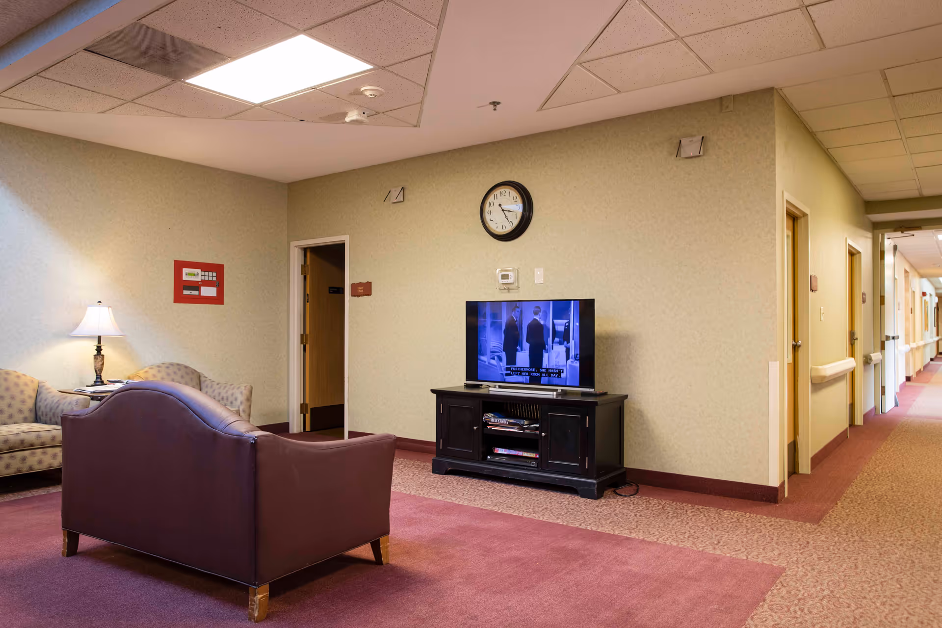 A common area in a senior living facility featuring a maroon couch facing a television on a black stand. There is a beige patterned loveseat with a lamp on a side table next to it. A clock is mounted on the wall above the television. The hallway with several doors extends to the right.