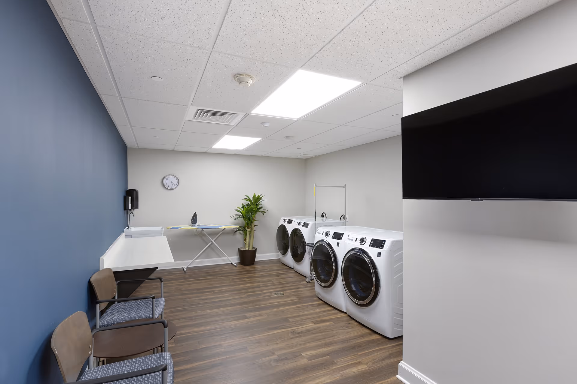 Laundry room with two front-loading washing machines and two dryers, an ironing board with an iron, a potted plant, a wall clock, two chairs with a small table between them, and a wall-mounted sink with a soap dispenser. The room has wood flooring, a blue accent wall, and a drop ceiling with fluorescent lights.