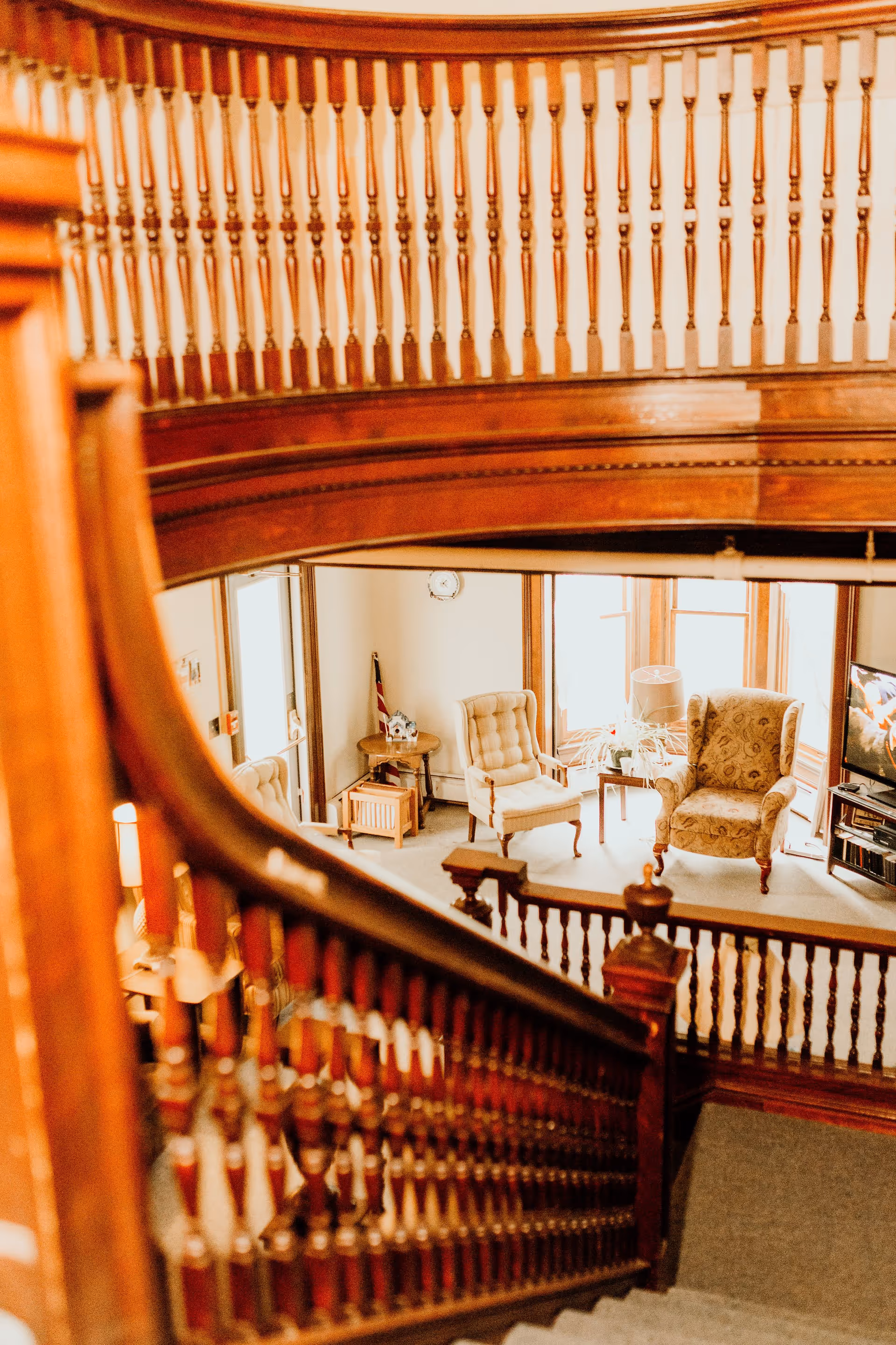 View down a wooden staircase with ornate banisters into a cozy living area featuring two upholstered armchairs, a small table with a lamp and plant, a television, and large windows letting in natural light.