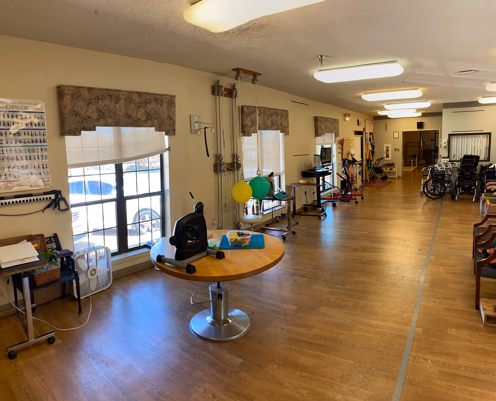 A long multipurpose activity and rehabilitation room with exercise equipment, wheelchairs, tables, and seating in a senior living facility.
