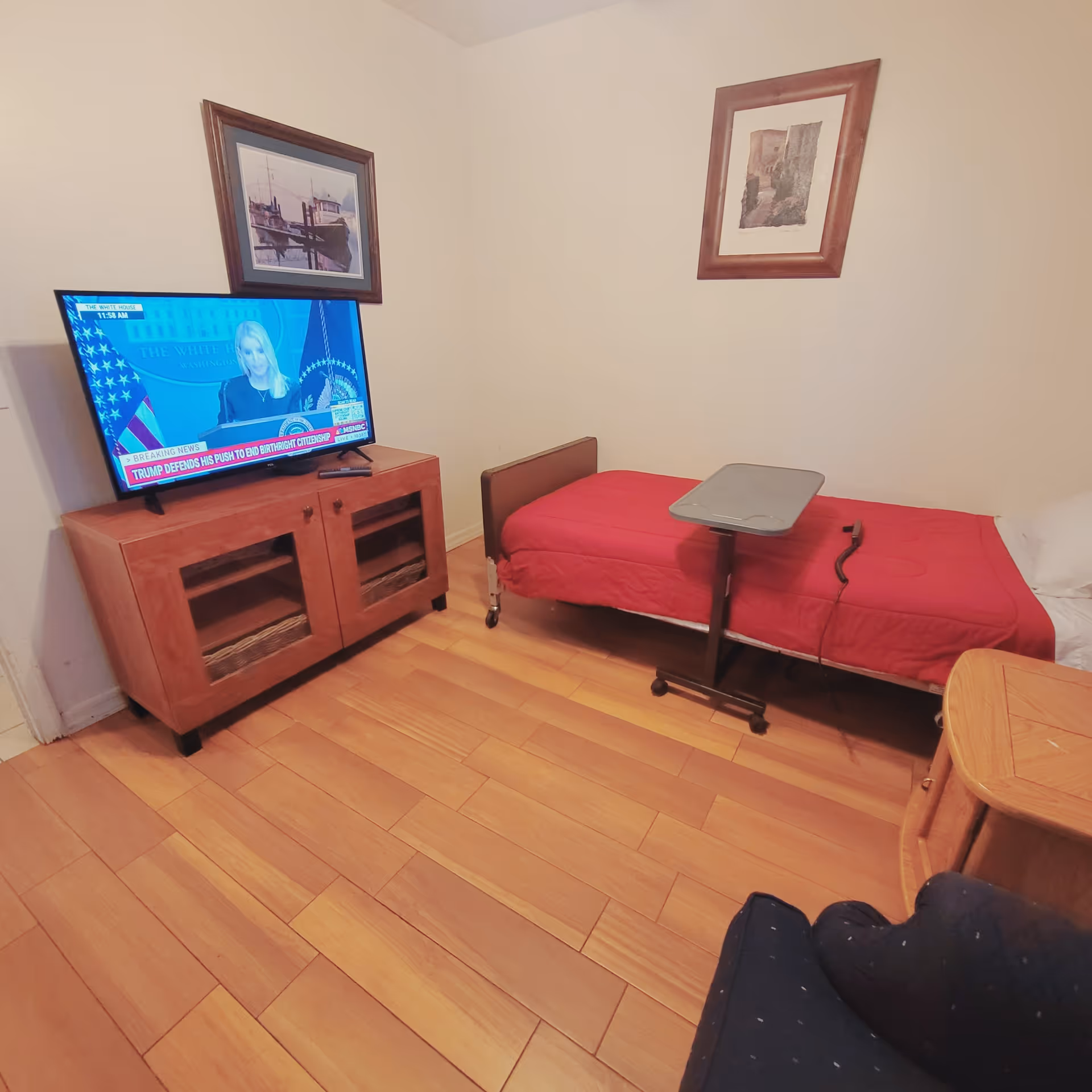 A small bedroom with a single bed covered with a red blanket, a movable overbed table, a wooden TV stand with a flat-screen TV showing a news broadcast, two framed pictures on the walls, and a wooden floor.