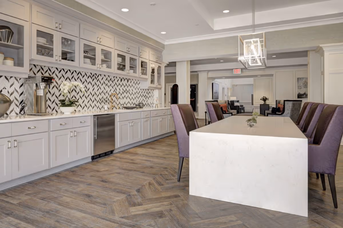 Modern communal kitchen and dining area with a long white island, purple chairs, and cabinetry with patterned backsplash.