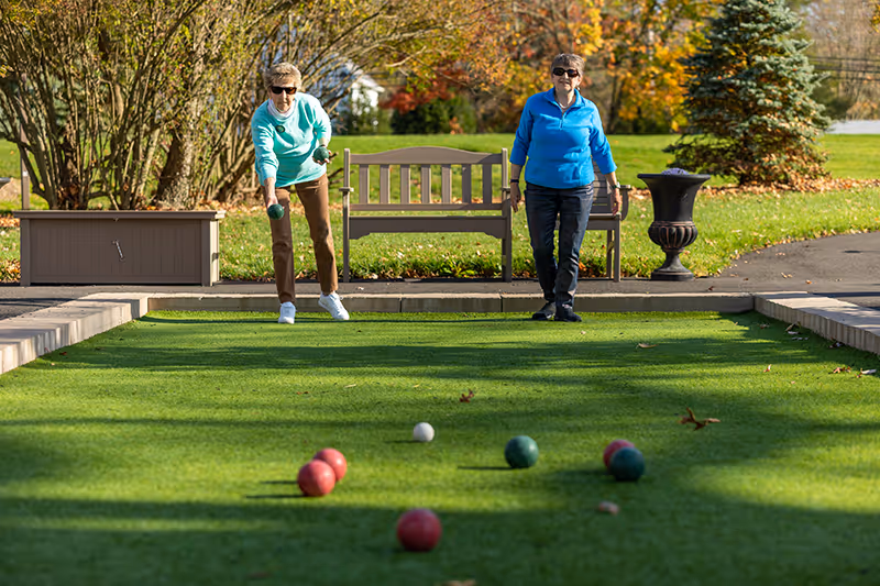 Two elderly women playing bocce ball on a green bocce court outdoors with trees and a bench in the background.