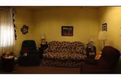 A living room with a patterned sofa in the center, flanked by two armchairs, one green and one maroon. There are two side tables with lamps on either side of the sofa, a window with curtains on the left, and framed pictures on the walls.