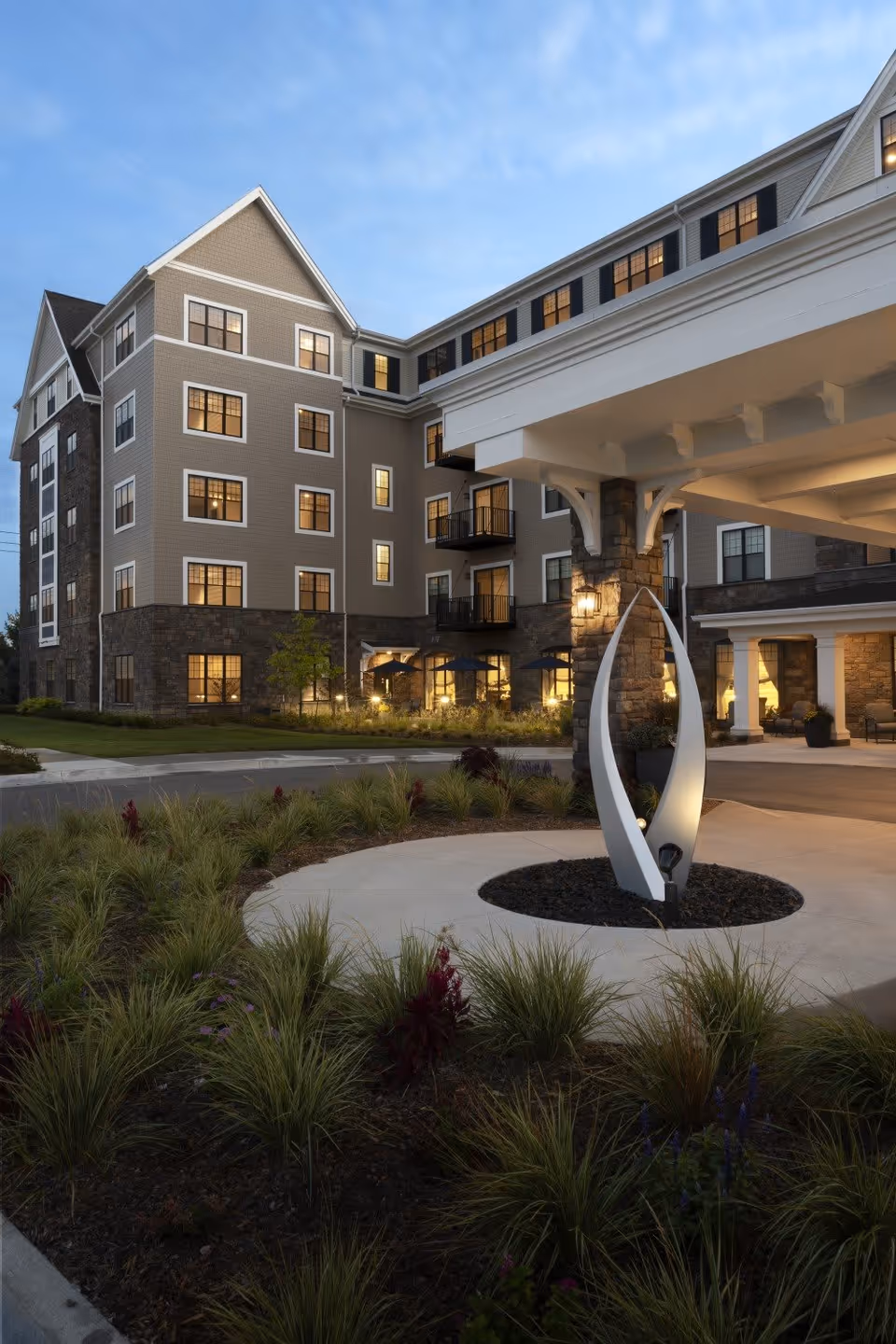 Exterior view of a multi-story senior living facility building at dusk with illuminated windows, landscaped garden with ornamental grasses and flowers, and a modern white sculpture near the entrance canopy.