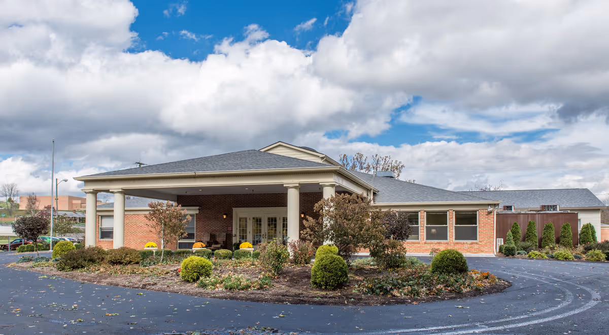 Exterior view of Lindside Healthcare Center, a single-story brick building with a covered entrance supported by white columns. The surrounding area features landscaped bushes and small trees under a partly cloudy sky.