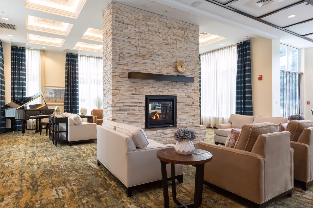 A spacious and well-lit living room area featuring a central stone fireplace with a modern black mantel. Surrounding the fireplace are beige and light brown upholstered armchairs and sofas arranged for conversation. Large windows with sheer white curtains and dark blue striped drapes allow natural light to fill the room. A grand piano is visible in the background, adding an elegant touch to the space. The ceiling has recessed lighting and decorative coffered panels.