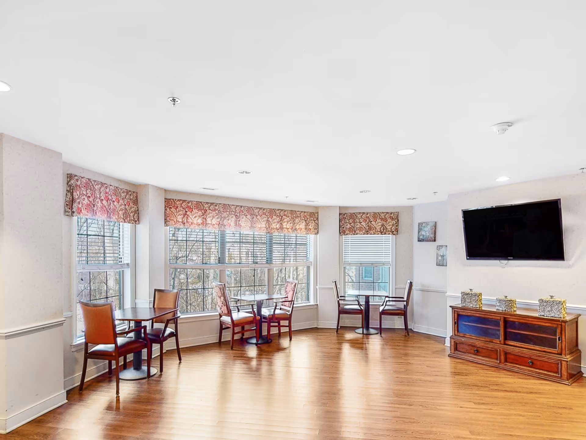A bright common area with large windows covered by floral valances, three small round tables each with two chairs, a wooden cabinet with decorative items on top, and a wall-mounted flat screen TV.