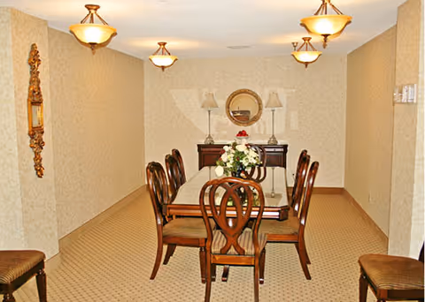 A formal dining room with a rectangular wooden table surrounded by six matching wooden chairs with cushioned seats. The room has beige patterned wallpaper and carpet, three ceiling light fixtures, a sideboard against the far wall with two table lamps and a round mirror above it, and a floral centerpiece on the table.