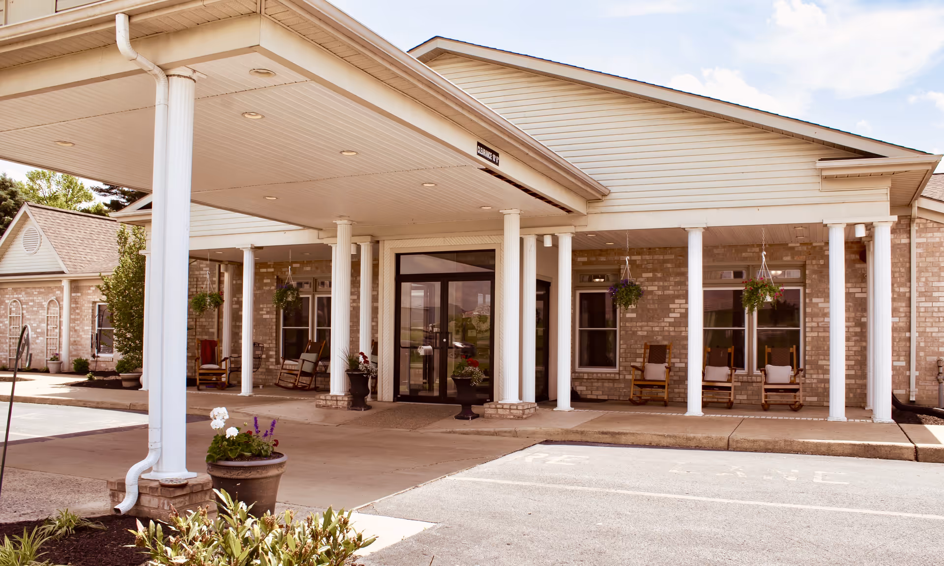 Entrance of a senior living facility with a covered driveway supported by white columns. The building has light-colored brick walls and several rocking chairs on the porch. There are hanging flower pots and potted plants near the entrance.