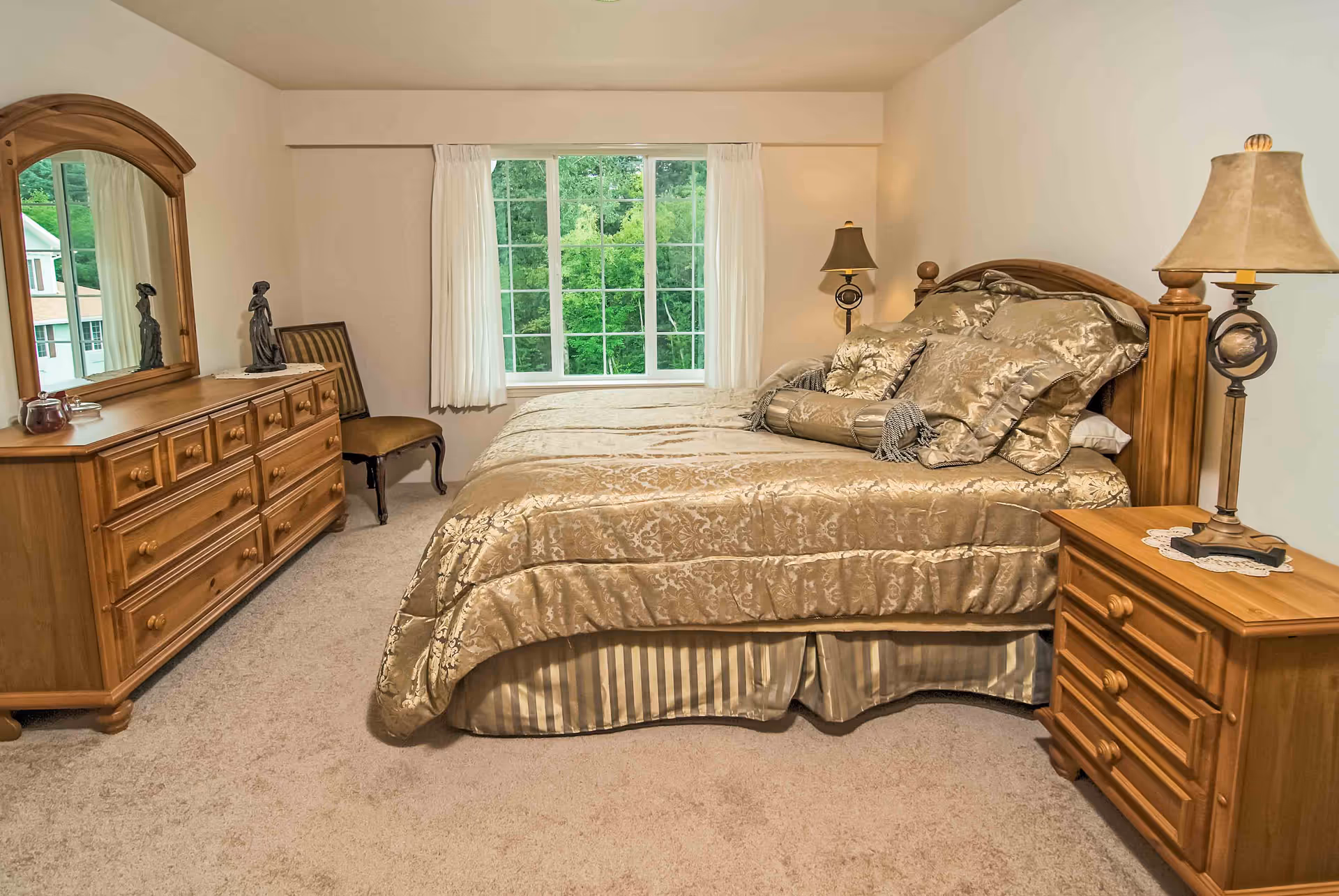 Well-furnished bedroom with a large bed, wooden dresser and nightstand, lamps, and a window overlooking greenery.