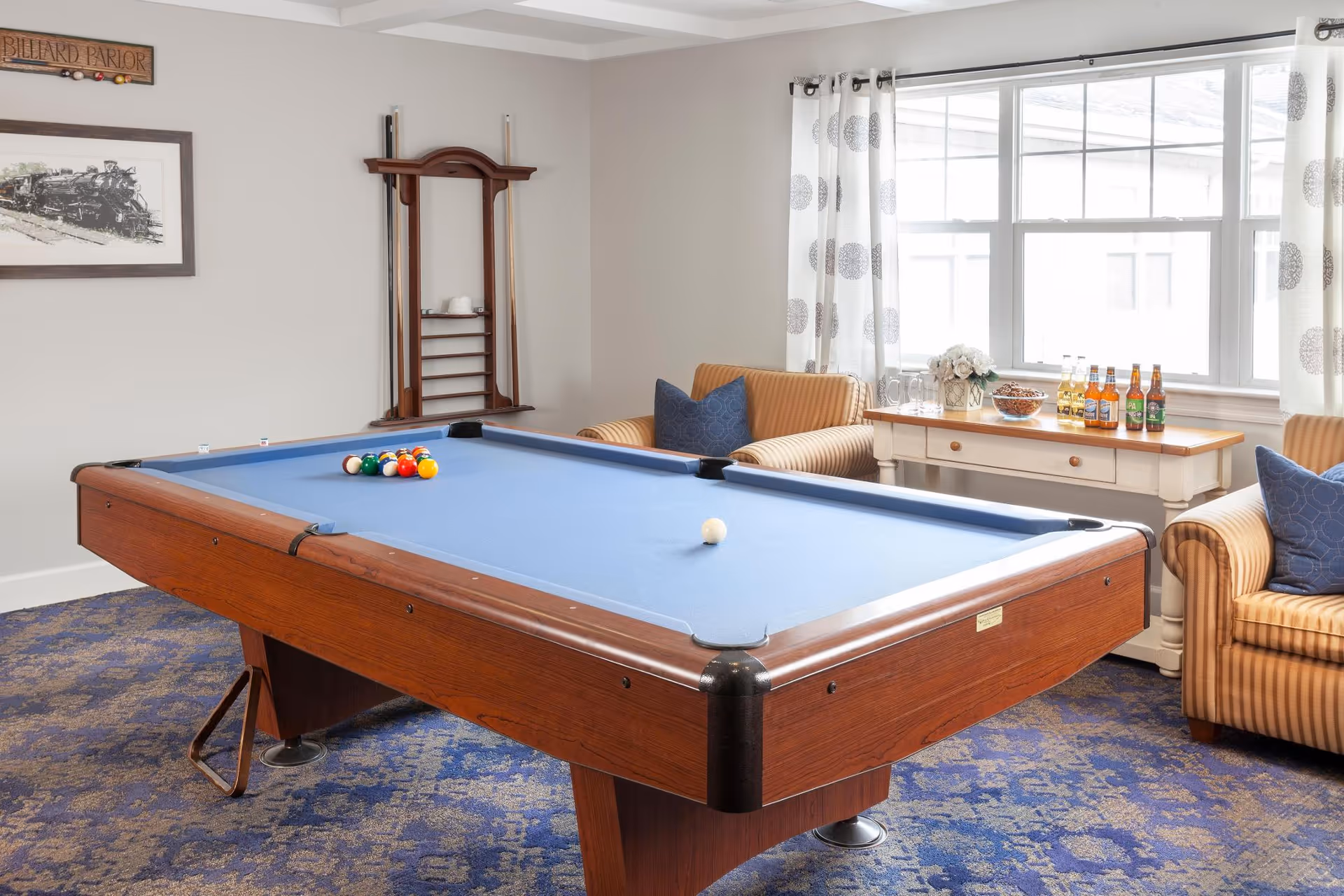 A bright room with a blue felt pool table in the center, a rack holding pool cues on the wall, two striped armchairs with blue cushions, a white side table with bottles and glasses, and a large window with patterned curtains letting in natural light.