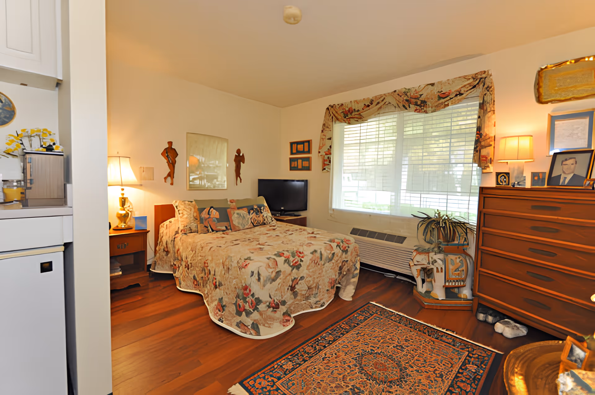 Sunlit cozy bedroom with a floral bedspread, wooden dresser, TV, and a window with blinds.