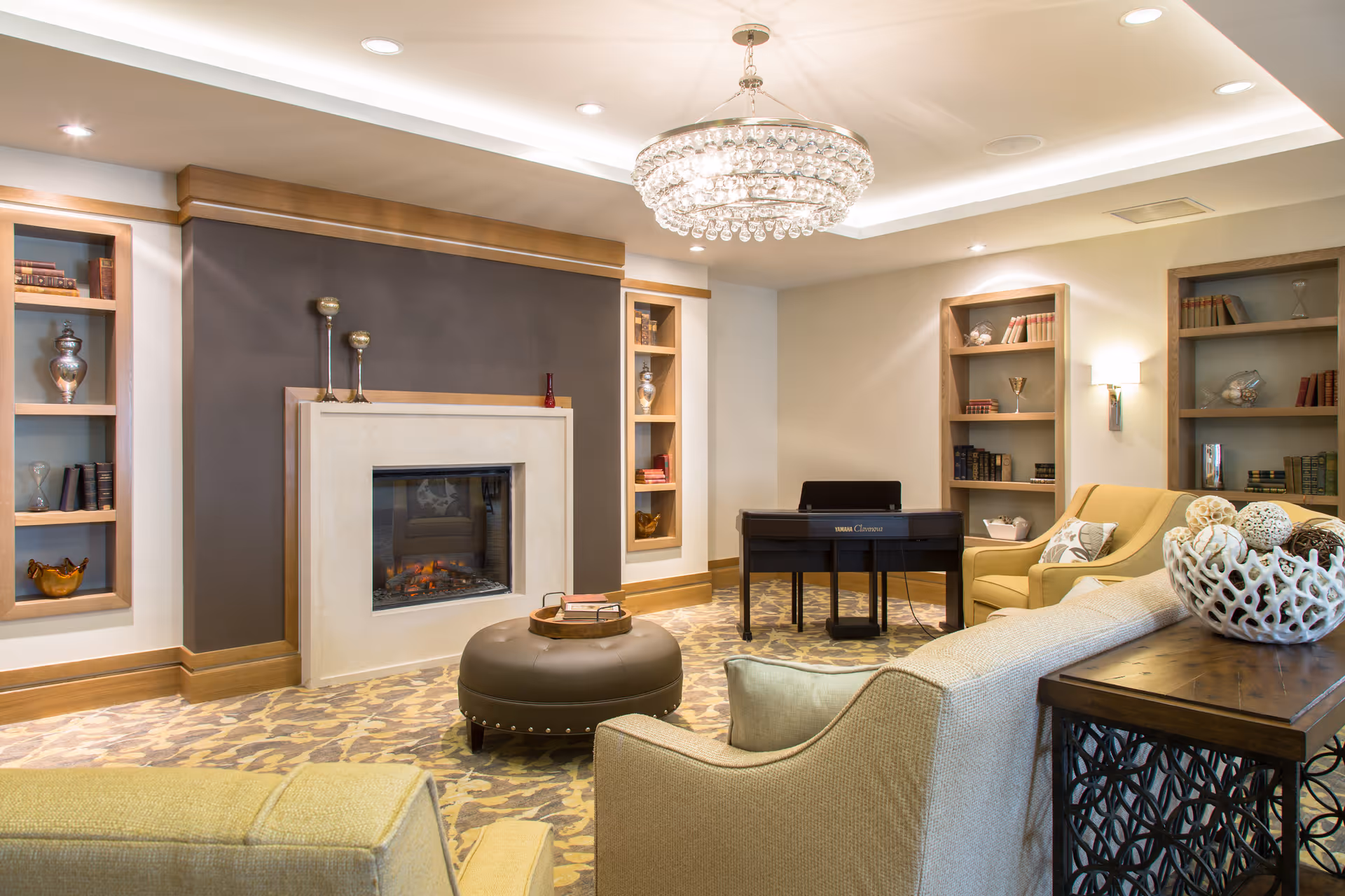 A cozy living room with a modern fireplace centered on a dark accent wall, flanked by built-in shelves holding decorative items and books. The room features comfortable beige armchairs and a sofa, a round brown ottoman with a tray on top, a wooden side table with a decorative bowl, and a Yamaha Clavinova digital piano. A large crystal chandelier hangs from the ceiling, illuminating the space.