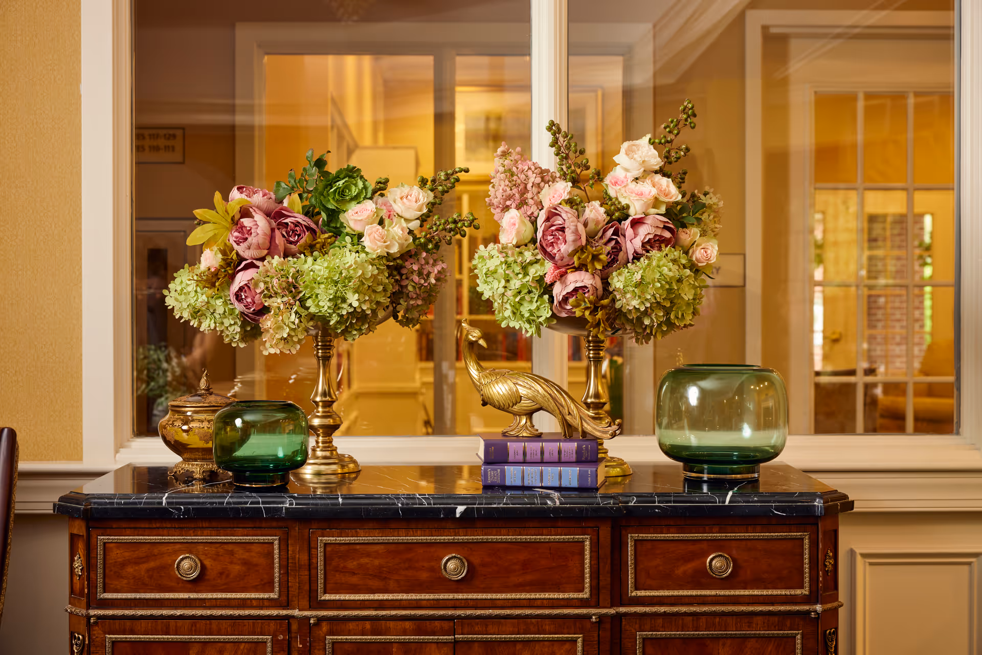 A polished wooden sideboard with a black marble top holds two ornate gold vases filled with lush floral arrangements of pink roses, green hydrangeas, and other flowers. Between the vases is a gold peacock figurine perched on a stack of three purple books. Two green glass decorative bowls flank the vases. Behind the sideboard is a large window with white trim, showing a warmly lit interior space with yellow walls and additional furnishings.