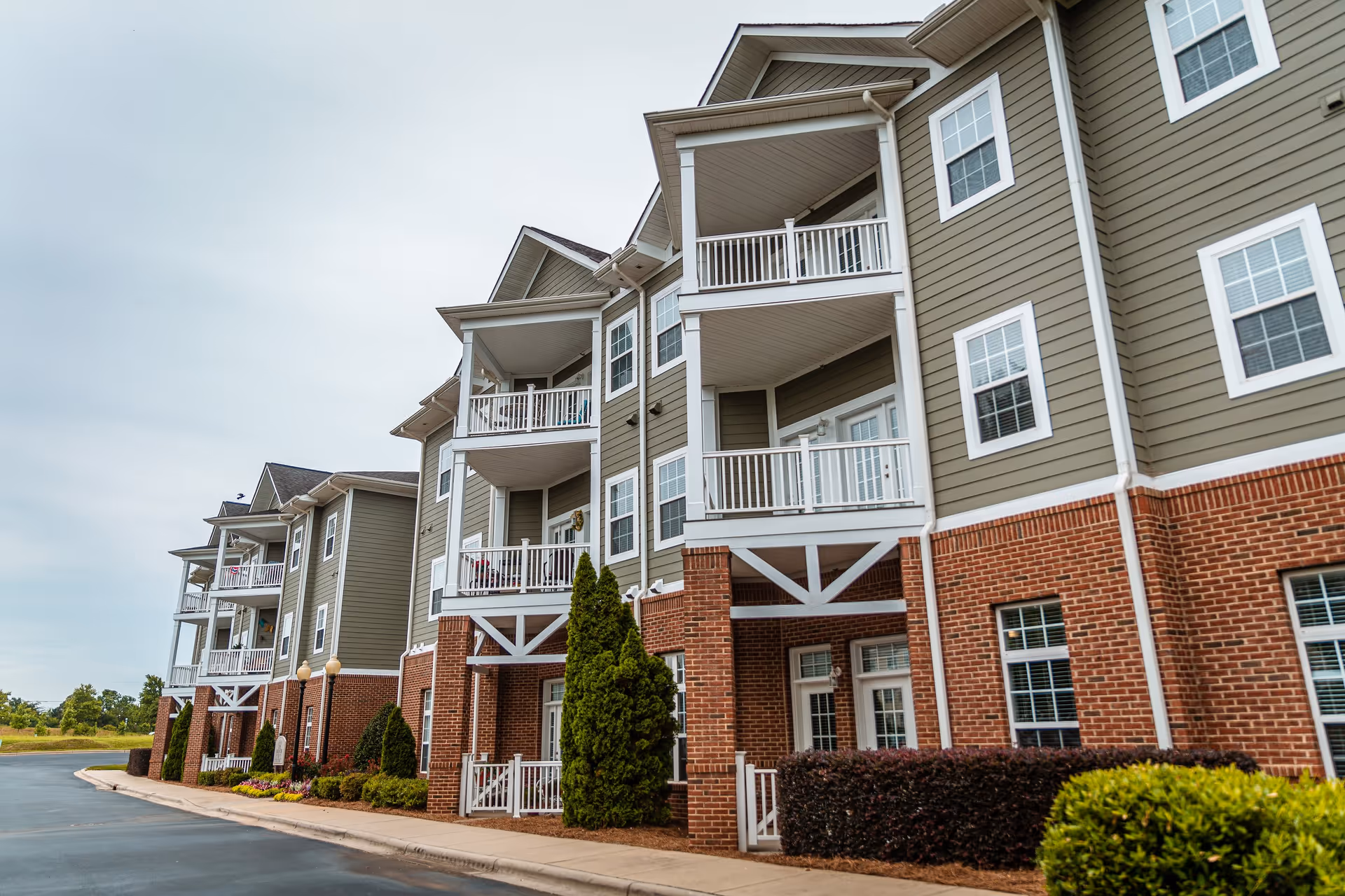 Exterior view of a multi-story residential building with balconies, brick and siding facade, surrounded by landscaped bushes and trees under a cloudy sky.