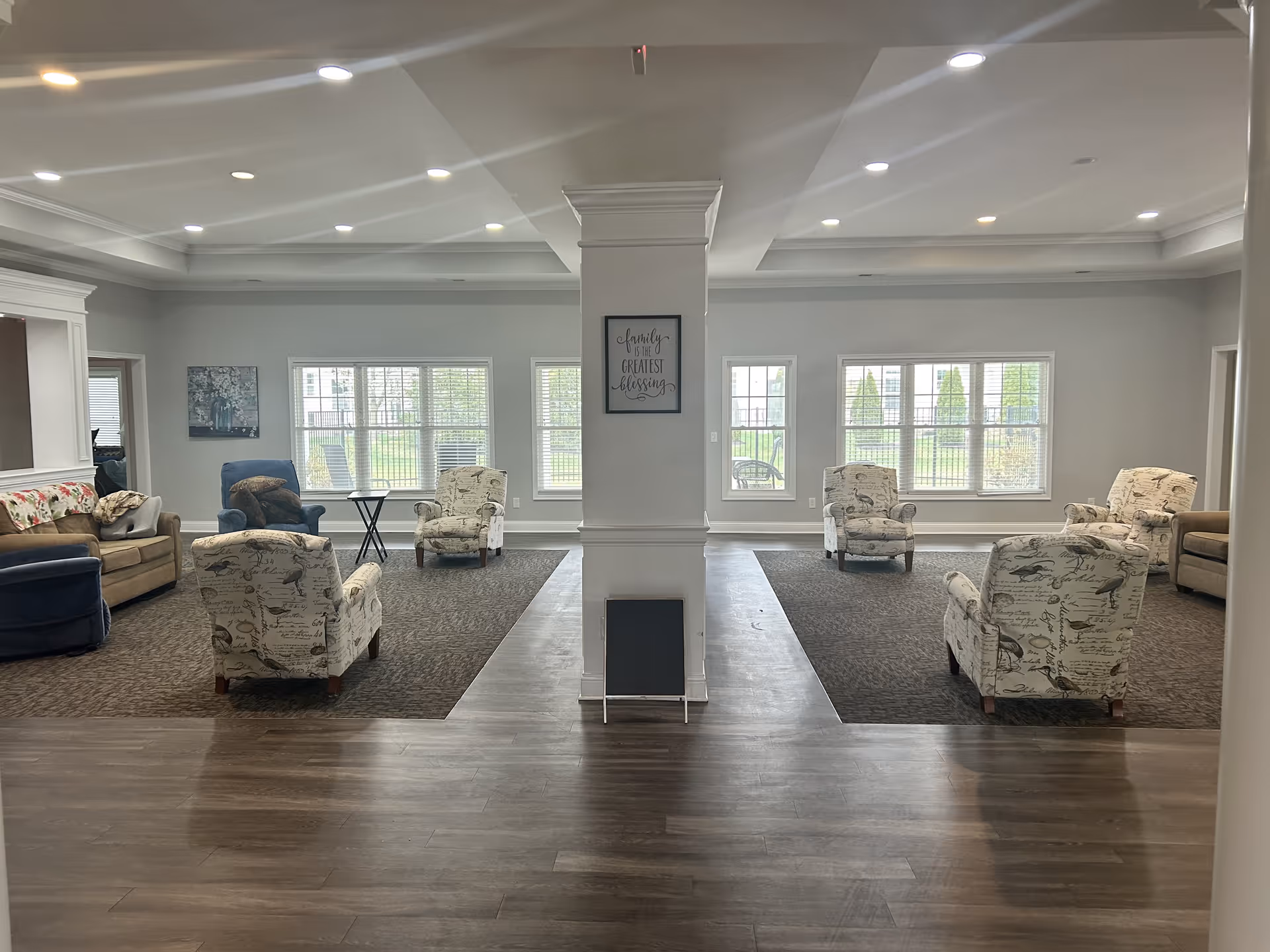 A spacious and well-lit living room area in a senior living facility with multiple armchairs and sofas arranged in two symmetrical seating areas on carpeted floors. Large windows allow natural light to fill the room, and a central pillar features a framed sign that reads 'Family is the greatest blessing.' The floor near the entrance is wooden, and the ceiling has recessed lighting.